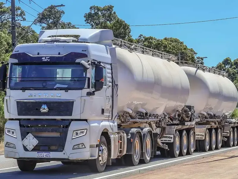 A large white tanker truck with multiple cylindrical containers is driving on a road. It is surrounded by greenery and trees, with power lines visible in the background. The truck has multiple wheels and visible license plates.